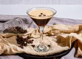 Un verre à martini rempli d'un Espresso Martini, avec une mousse crémeuse garnie de trois grains de café. En arrière-plan, un pot en verre renversé laisse échapper des grains de café sur une serviette beige, créant une ambiance chaleureuse et raffinée.