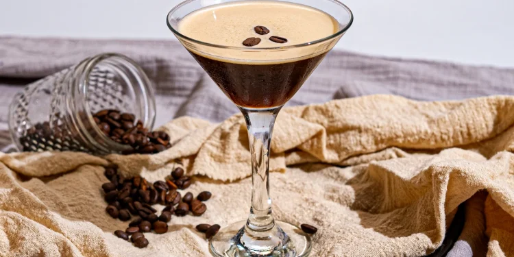 Un verre à martini rempli d'un Espresso Martini, avec une mousse crémeuse garnie de trois grains de café. En arrière-plan, un pot en verre renversé laisse échapper des grains de café sur une serviette beige, créant une ambiance chaleureuse et raffinée.