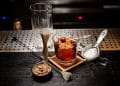 Un verre old-fashioned contenant un cocktail Vieux Carré, garni d’une cerise confite sur un pic décoratif et d’un gros glaçon. Autour du verre, un verre à mélange, un doseur en métal, une passoire à cocktail et un bol de fèves de cacao reposent sur un comptoir de bar élégant.