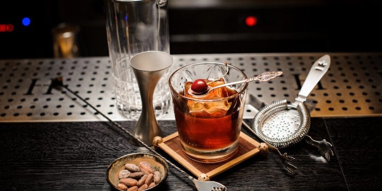 Un verre old-fashioned contenant un cocktail Vieux Carré, garni d’une cerise confite sur un pic décoratif et d’un gros glaçon. Autour du verre, un verre à mélange, un doseur en métal, une passoire à cocktail et un bol de fèves de cacao reposent sur un comptoir de bar élégant.