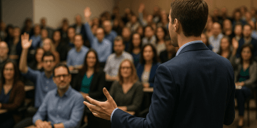 Conférencier brun en costume vu de dos, parlant avec les mains devant une salle pleine où plusieurs participants lèvent la main pour poser des questions.